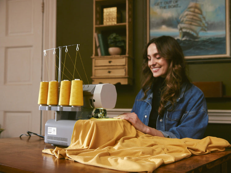 A smiling woman sews yellow fabric using a Singer Overlocker with yellow thread
