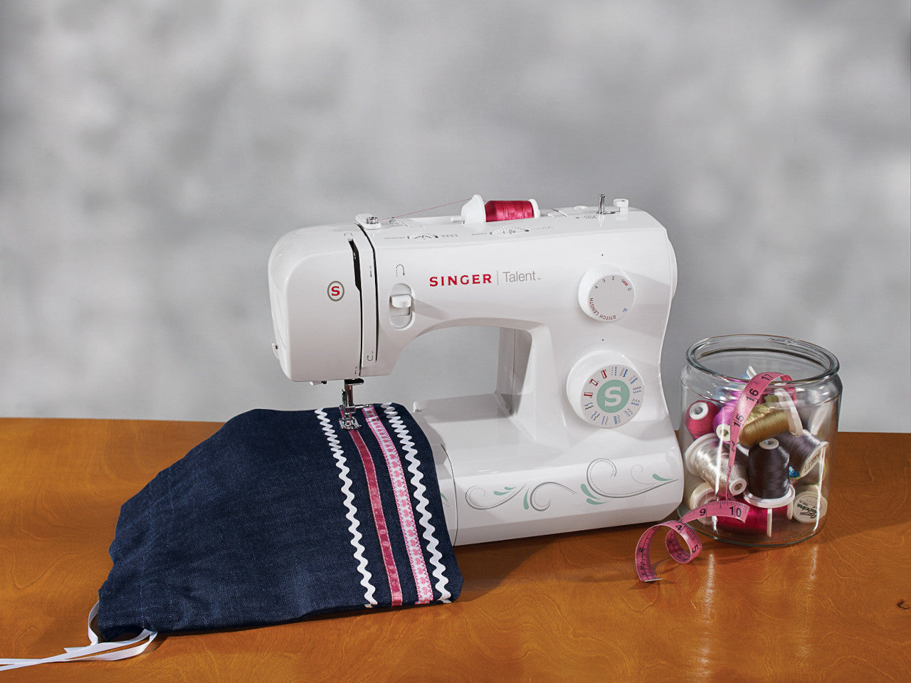 A white SINGER Talent 3321 sewing machine stitching a white trim on a dark denim pouch, next to a glass jar with different coloured threads and measuring tape.