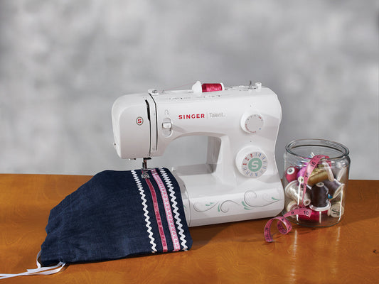 A white SINGER Talent 3321 sewing machine stitching a white trim on a dark denim pouch, next to a glass jar with different coloured threads and measuring tape.