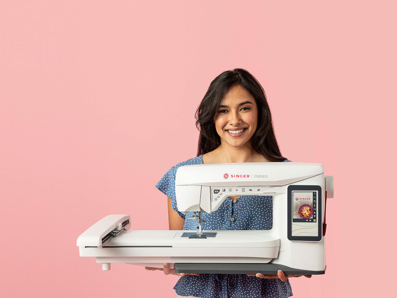 A smiling woman with her hands extended holding the SINGER EM9305 embroidery machine against a pink background.