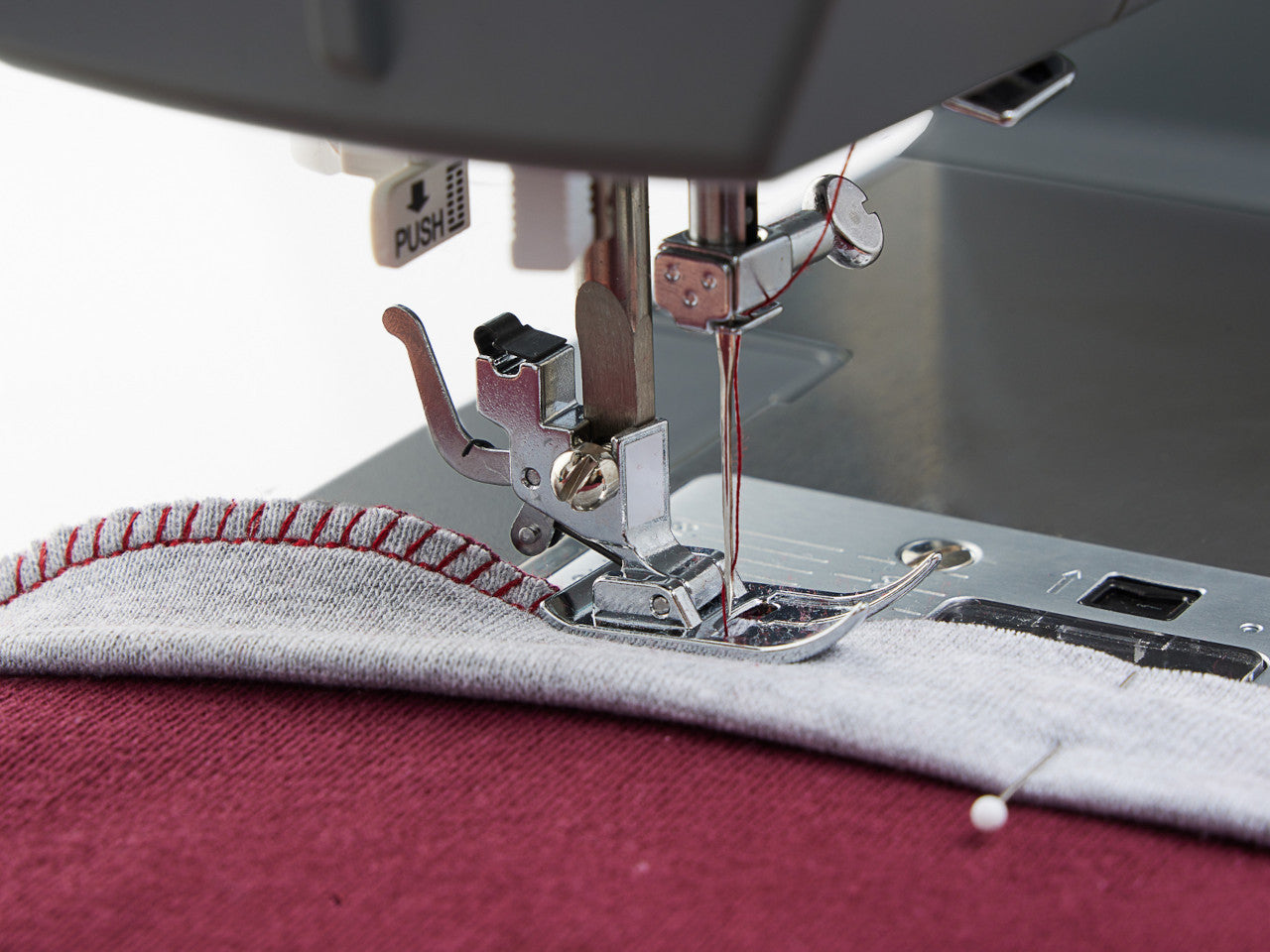 Close up of the presser foot stitching a maroon and light grey jersey knit fabric on a SINGER heavy duty 4423 sewing machine capable of 97 stitches.
