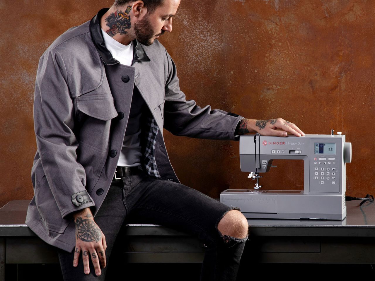 A tattooed man with torn jeans and a grey coat sits with one leg on a stainless steel table with one hand on the top of a Singer HD6805C sewing machine.