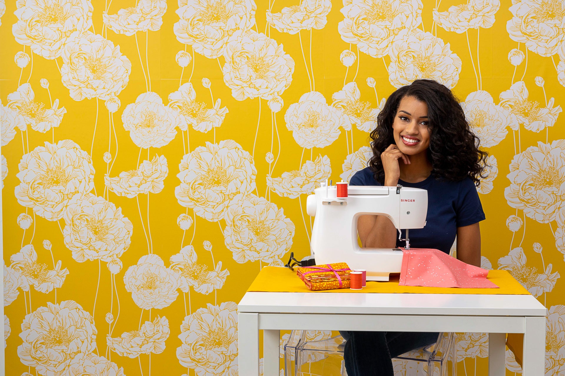 A smiling woman rests her chin on her hand while sitting behind a white table, which has a Singer sewing machine, stitching a salmon-coloured fabric. The wallpaper is bright yellow with a white floral pattern.