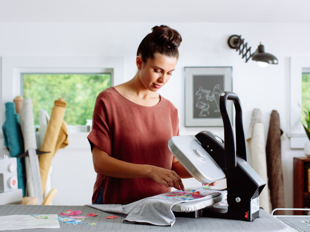 A woman with her hair tied in a bun, positions a shirt in the SINGER Professional Steam Press with rolls of fabric in the background of the sewing room.
