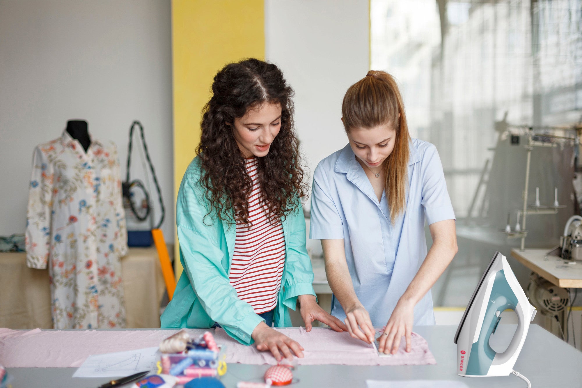 Two young women draw a pattern line on pink fabric on a sewing table with a Singer steam iron close by