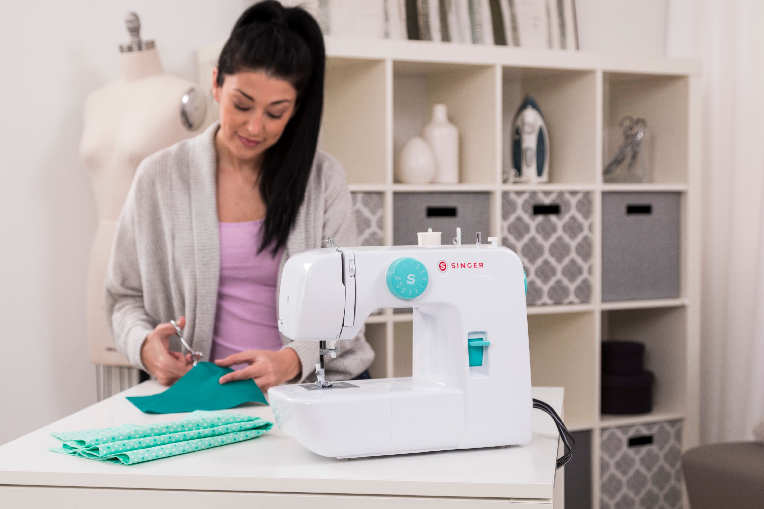A lady cuts green fabric with a pair of scissors with a Singer sewing machine on the table top