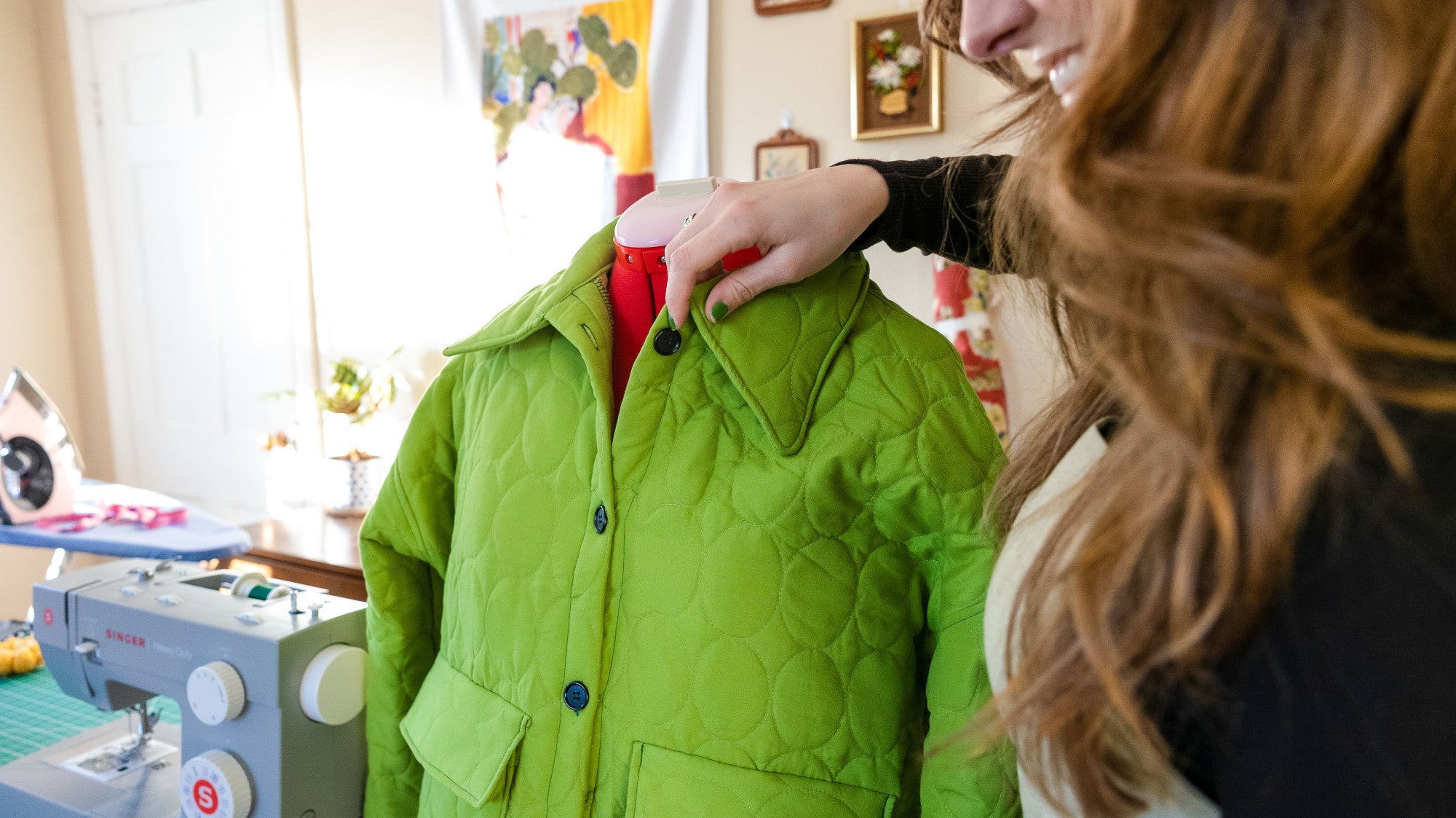 A smiling woman holds the collar of a green quilted jacket which is mounted to a Singer dress form, with a grey Singer heavy duty sewing machine on the table alongside her.