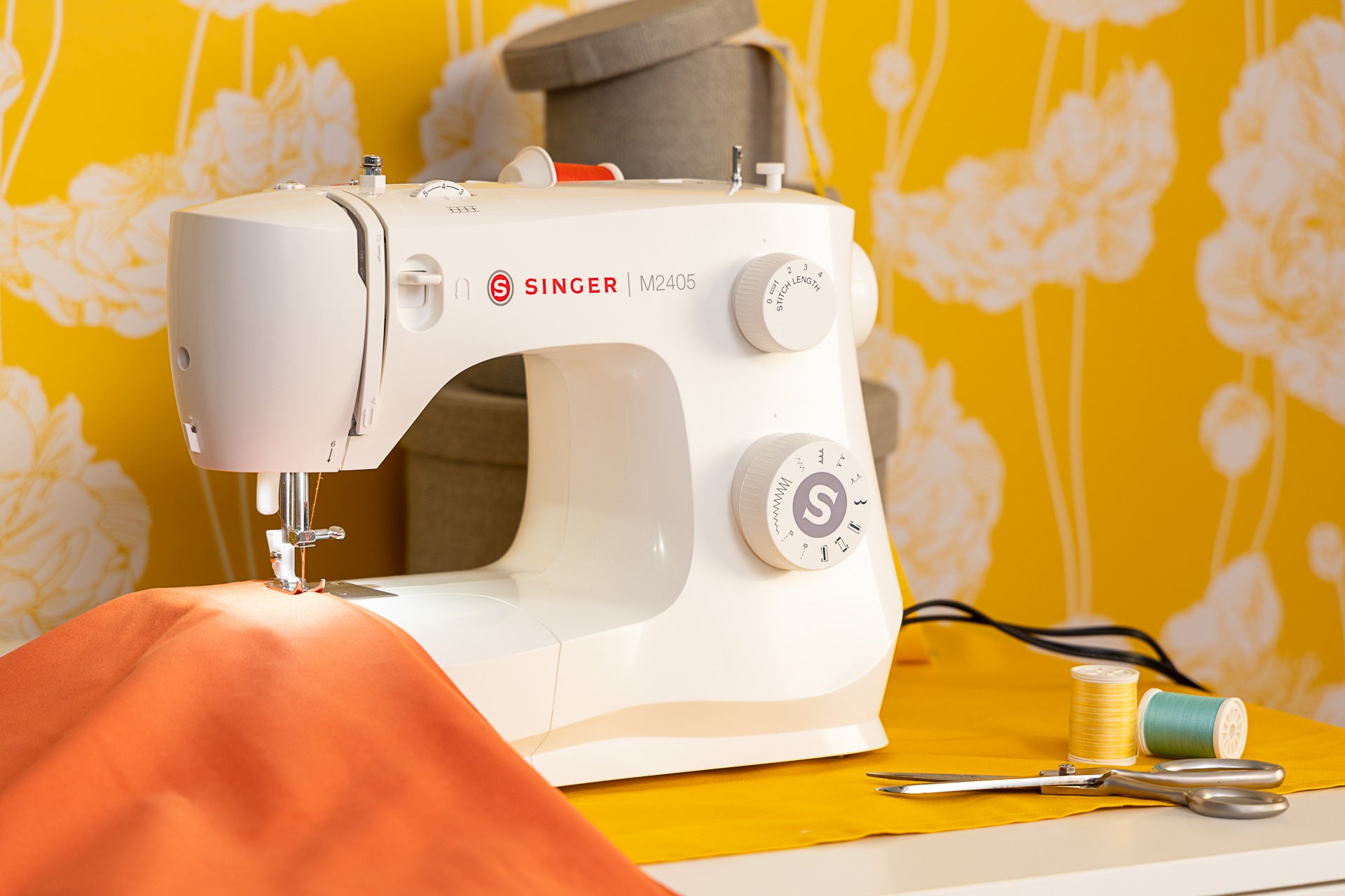 A white SINGER M2405 sewing machine with orange fabric ready to sew against a vibrant yellow wall with white floral patterns